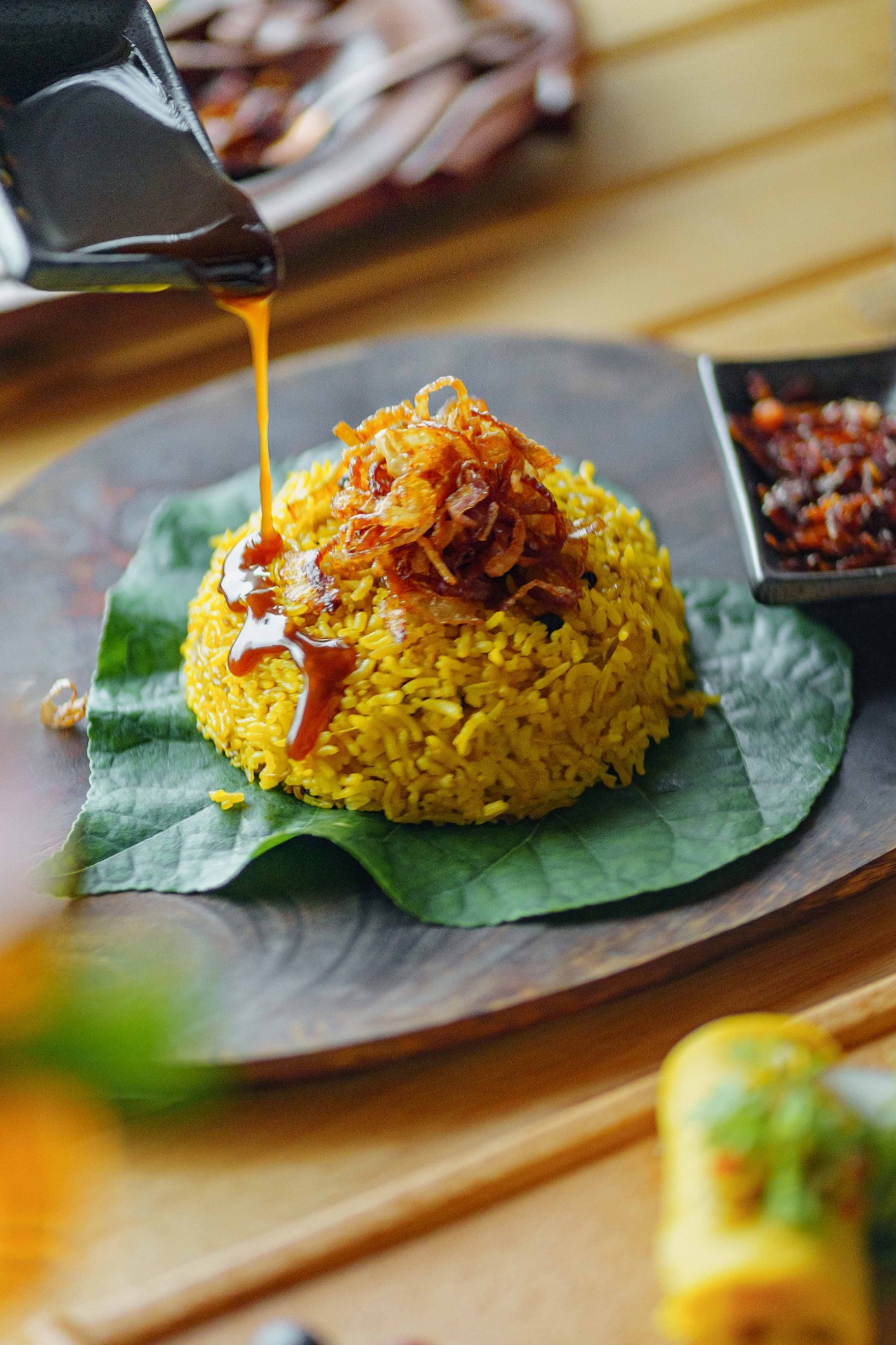 Sauce being poured over a rice dish topped with fried onions and served on a leaf